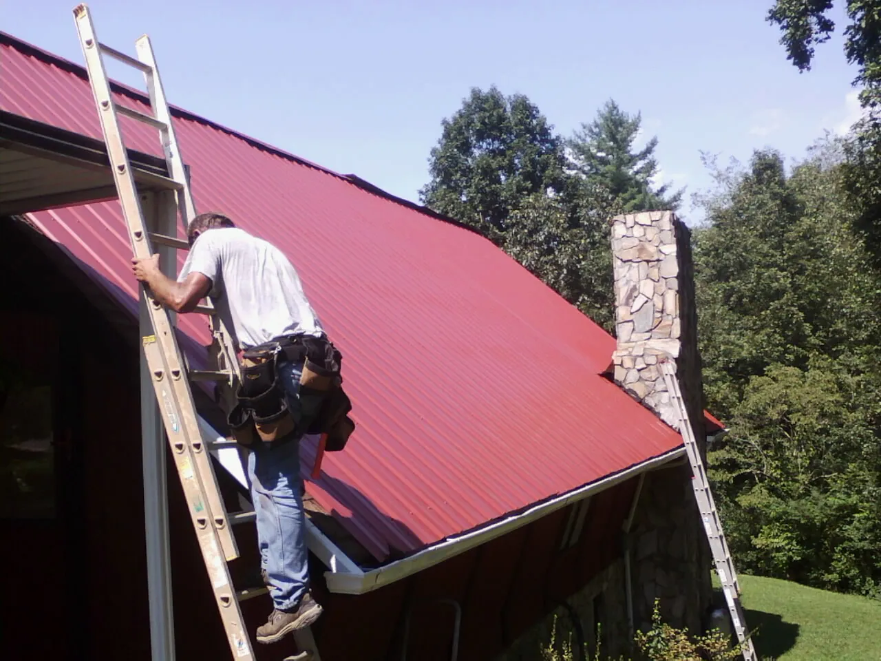 Roofer on a ladder beside a red metal roof and stone chimney — Soffit & Fascia Repair by Roofing Brothers in East Walpole